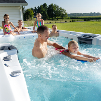 A dad helping a daughter on a kickboard swim against the propulsion in a swim spa with the rest of the family watching.