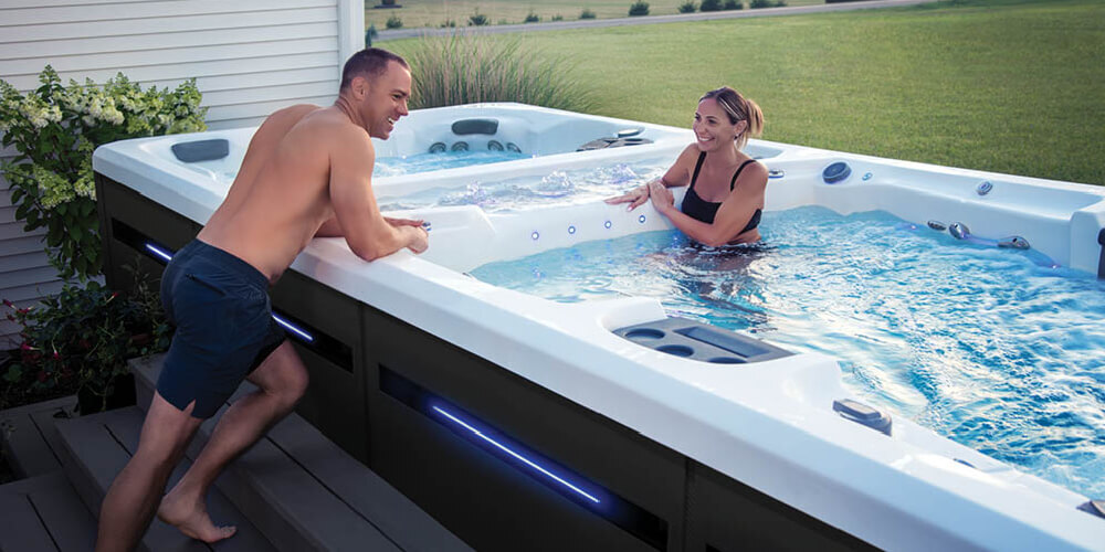 Man leaning on the side of a swim spa talking to a woman who is in the water. 